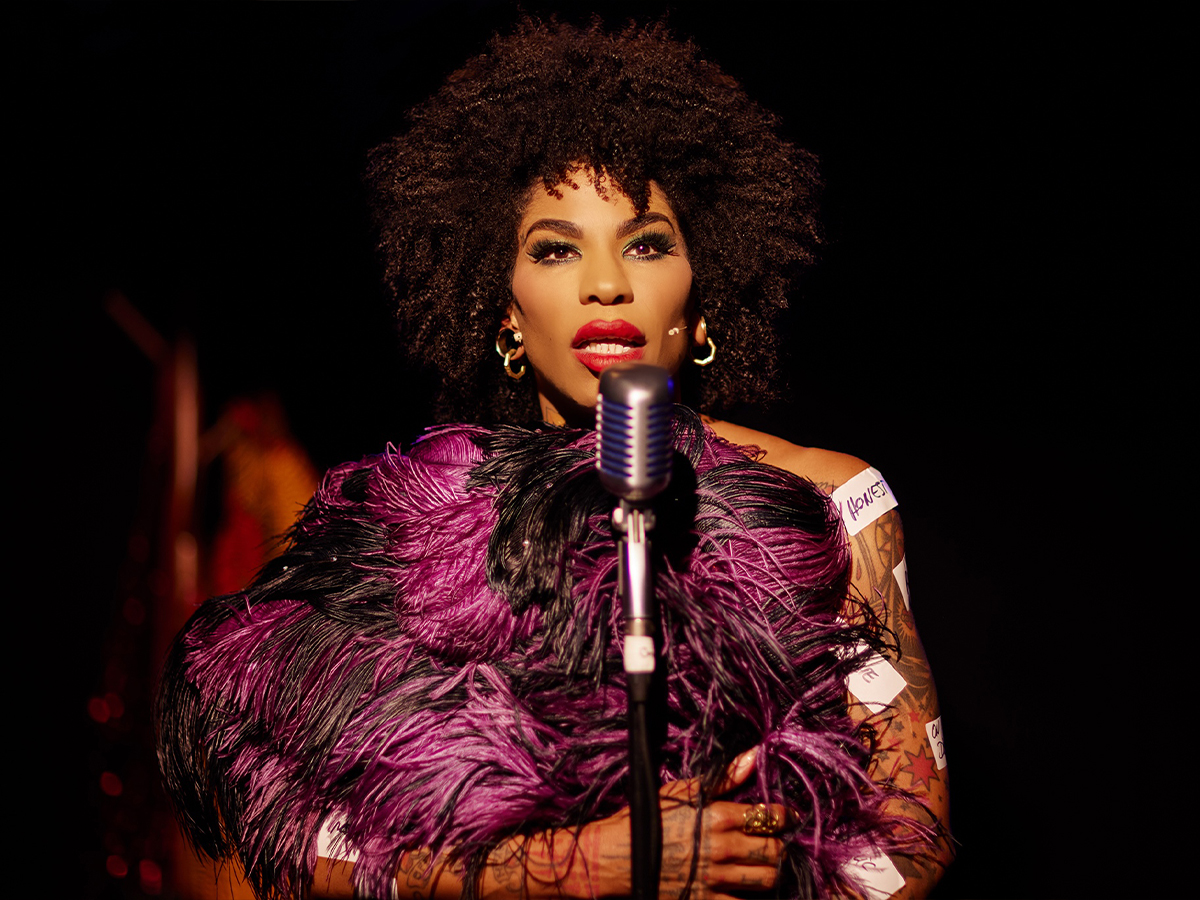 Image shows a black artist on stage in front of a microphone. They are holding feathers and have sticky labels attached to them.