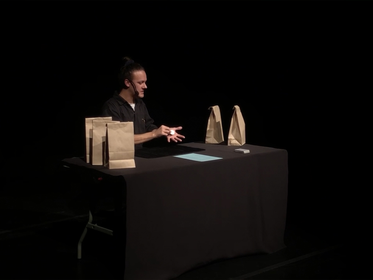 A performer sits at a desk with multiple brown paper bags.