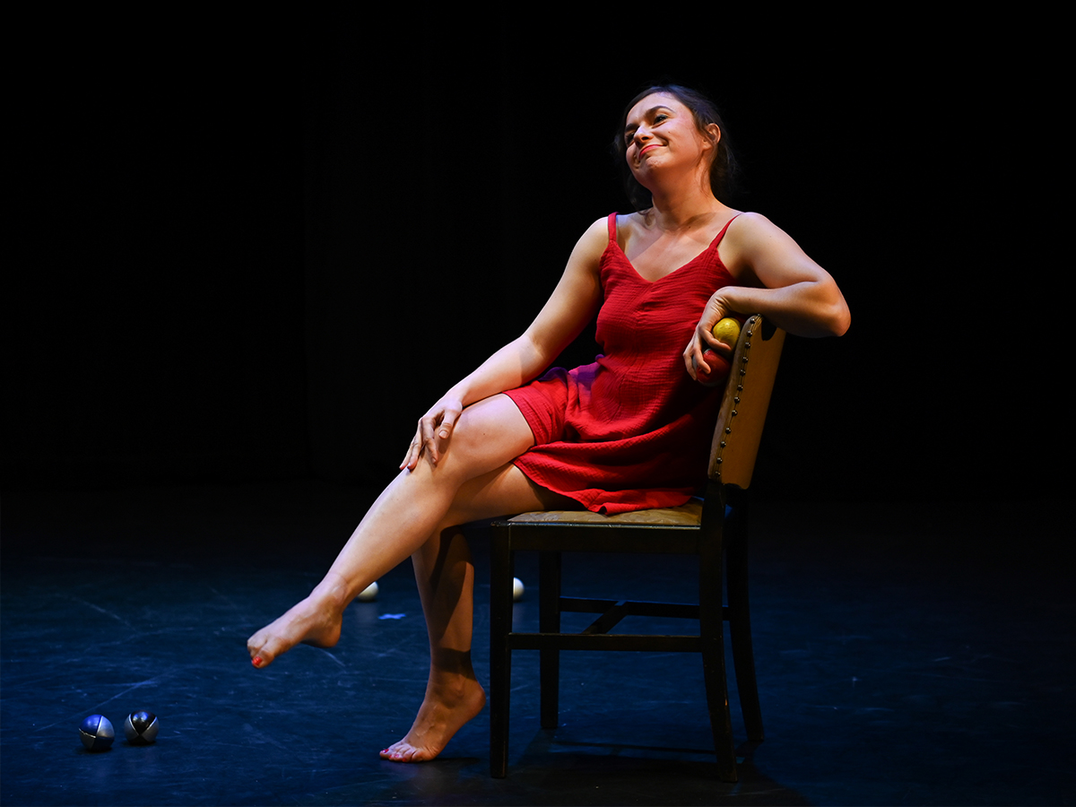 Image shows a performer in a red dress, sat on a chair, holding juggling balls.