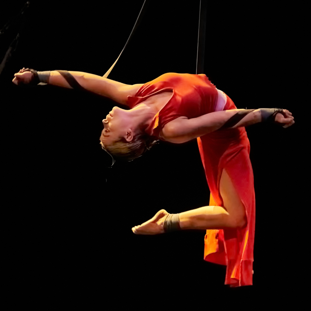 A straps aerialist hanging in a back bend with the straps wrapped around their arms. The aerialist is wearing a red dress.