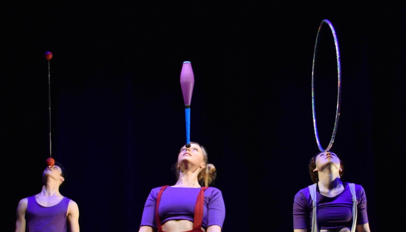 Three jugglers wearing purple, each balancing a prop on their head. A juggler balancing a staff on the left. A juggler balancing a club in the middle. A juggler balancing a hula hoop on the right.