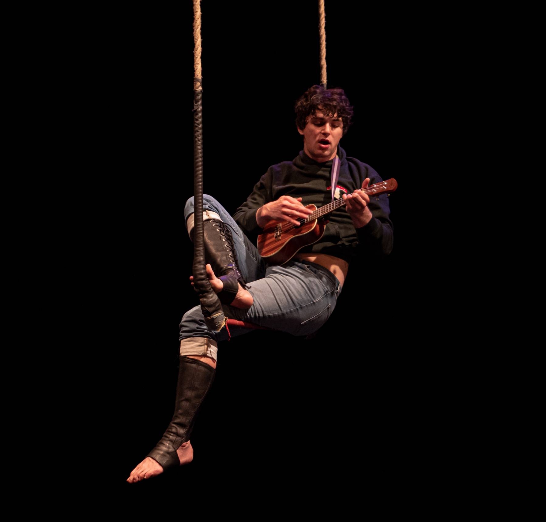 A man with a ukulele sits on a trapeze high against a black background
