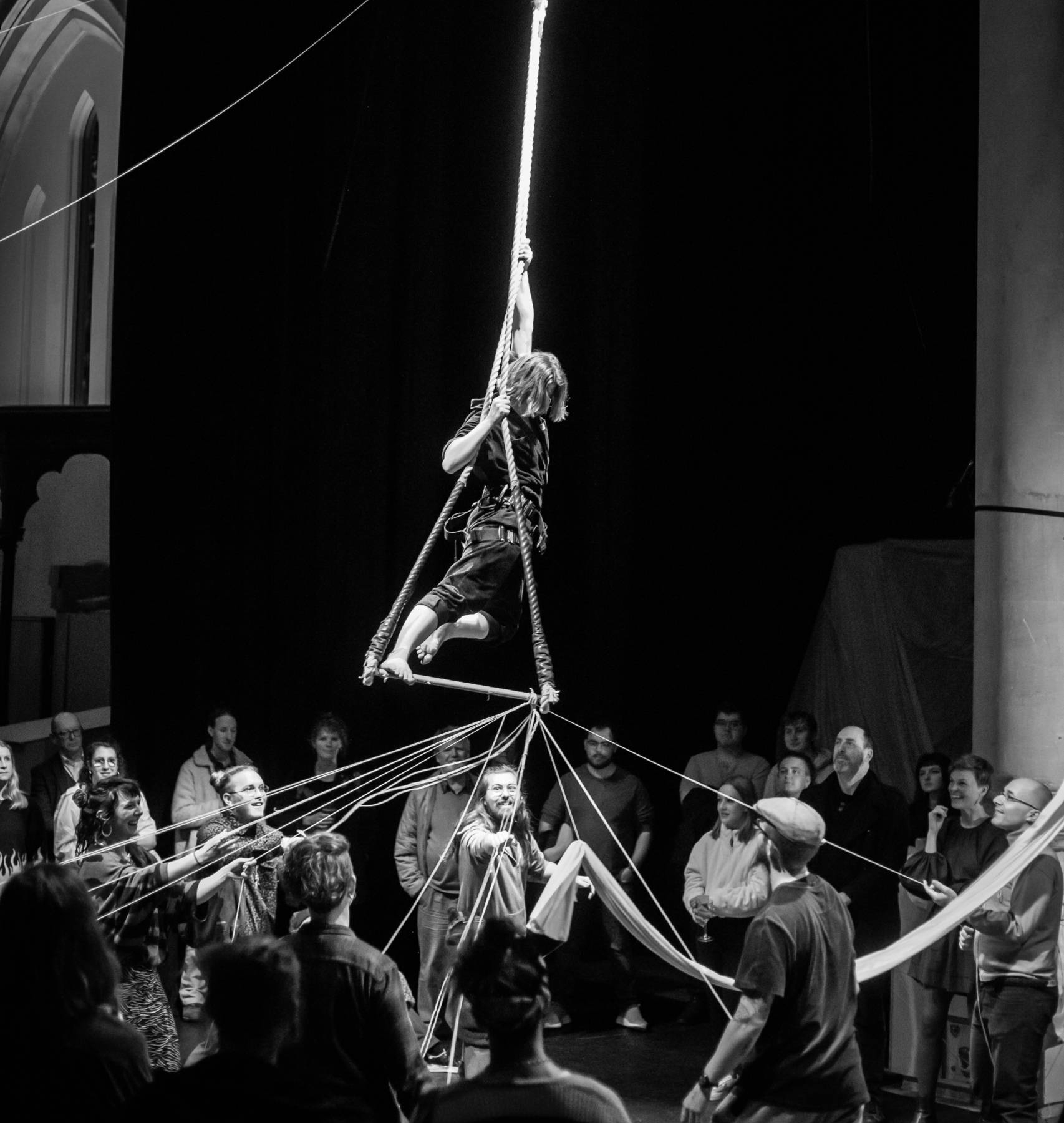 a black and white photo of a person on trapeze being pulled by many pieces of string by the audience