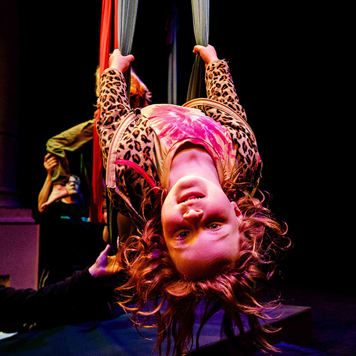 A young circus student hangs upside down in aerial silks, smiling at the camera, with an instructor and another student visible in the background